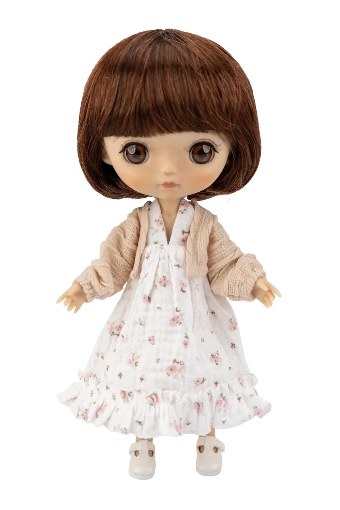 A doll with brown hair and large eyes wearing a white floral dress with a beige cardigan and white shoes, shown against a plain white background.