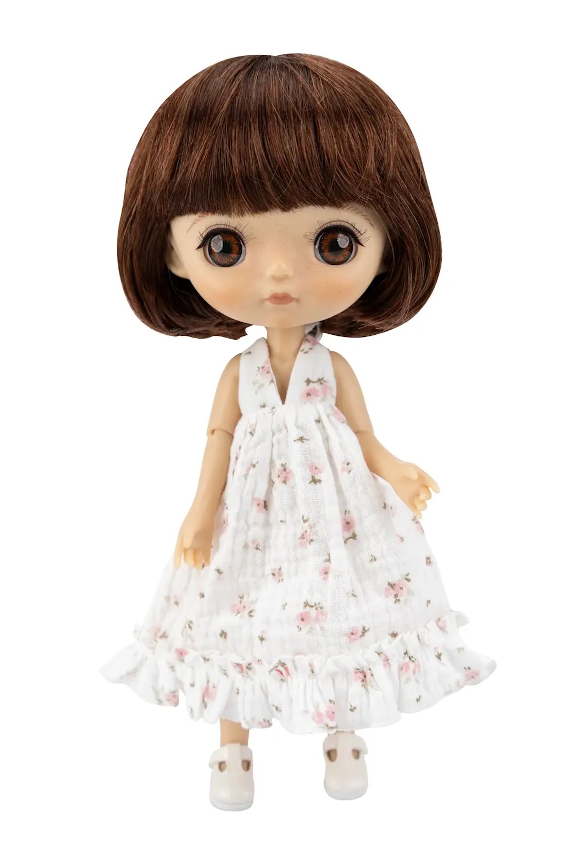 A doll with brown hair and large eyes wearing a white sleeveless dress with a pink floral pattern and white shoes, isolated on a white background.