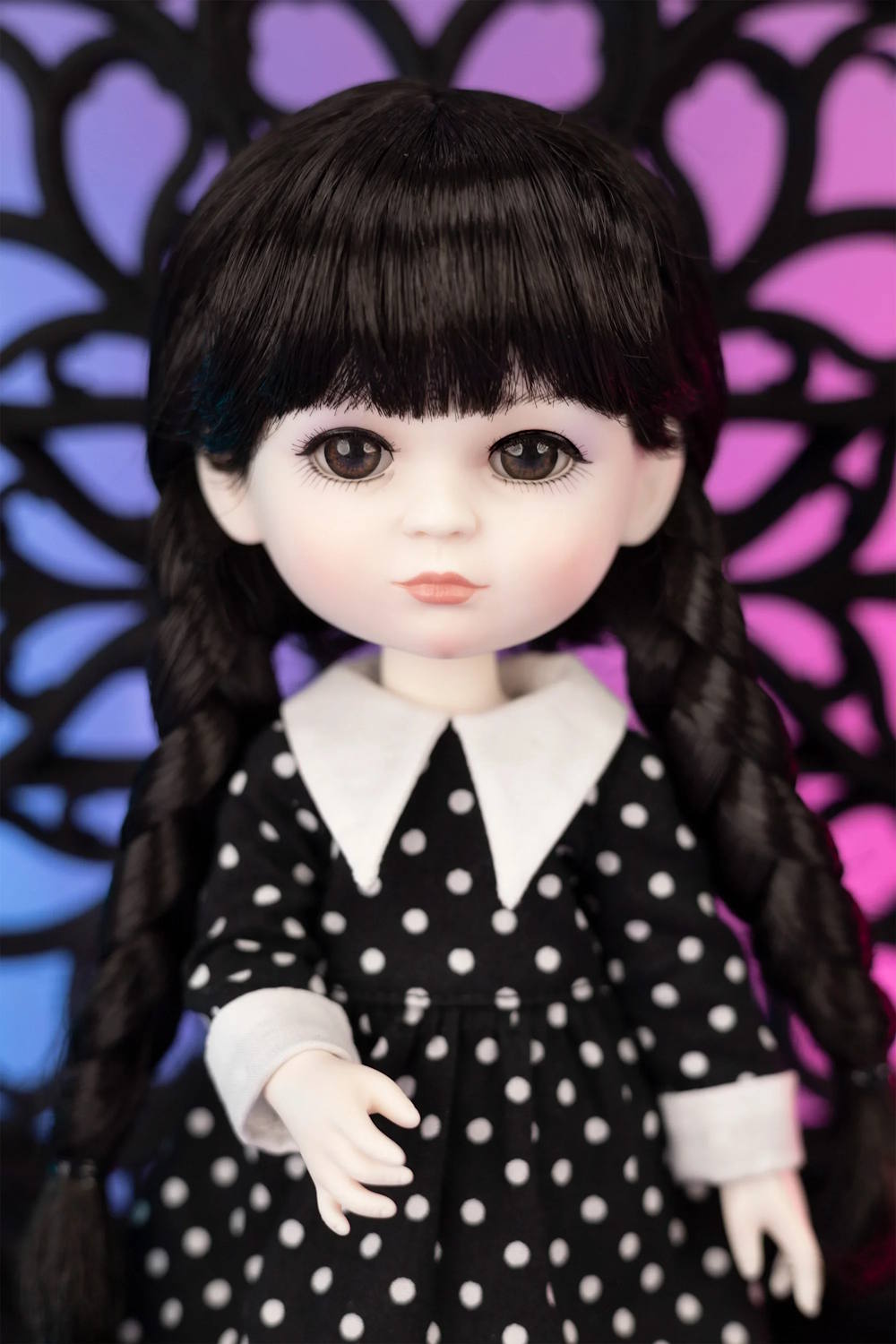 Doll with black hair in braids wearing a black dress with white polka dots against a colorful abstract background.