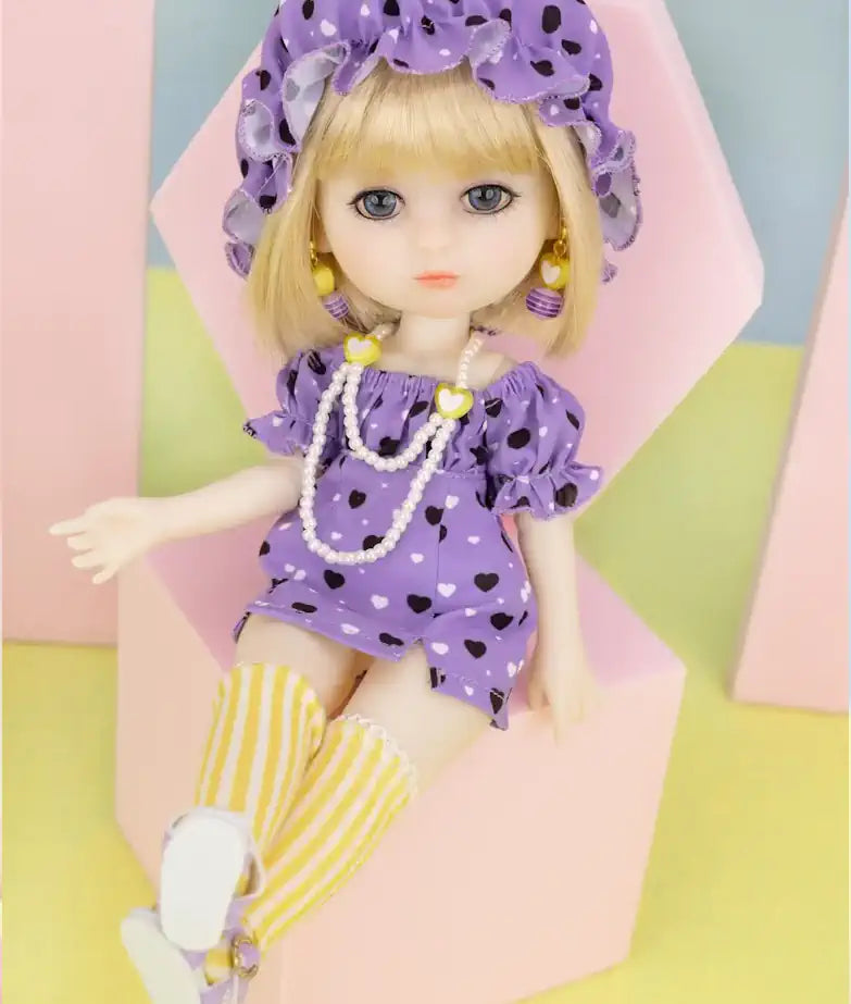 Luna Lily Doll Image Photo: A Luna Lily Doll dressed in a purple outfit adorned with hearts, wearing a matching hat and accessories. The doll has blonde hair and is seated on a pink geometric background, showcasing charming striped socks.