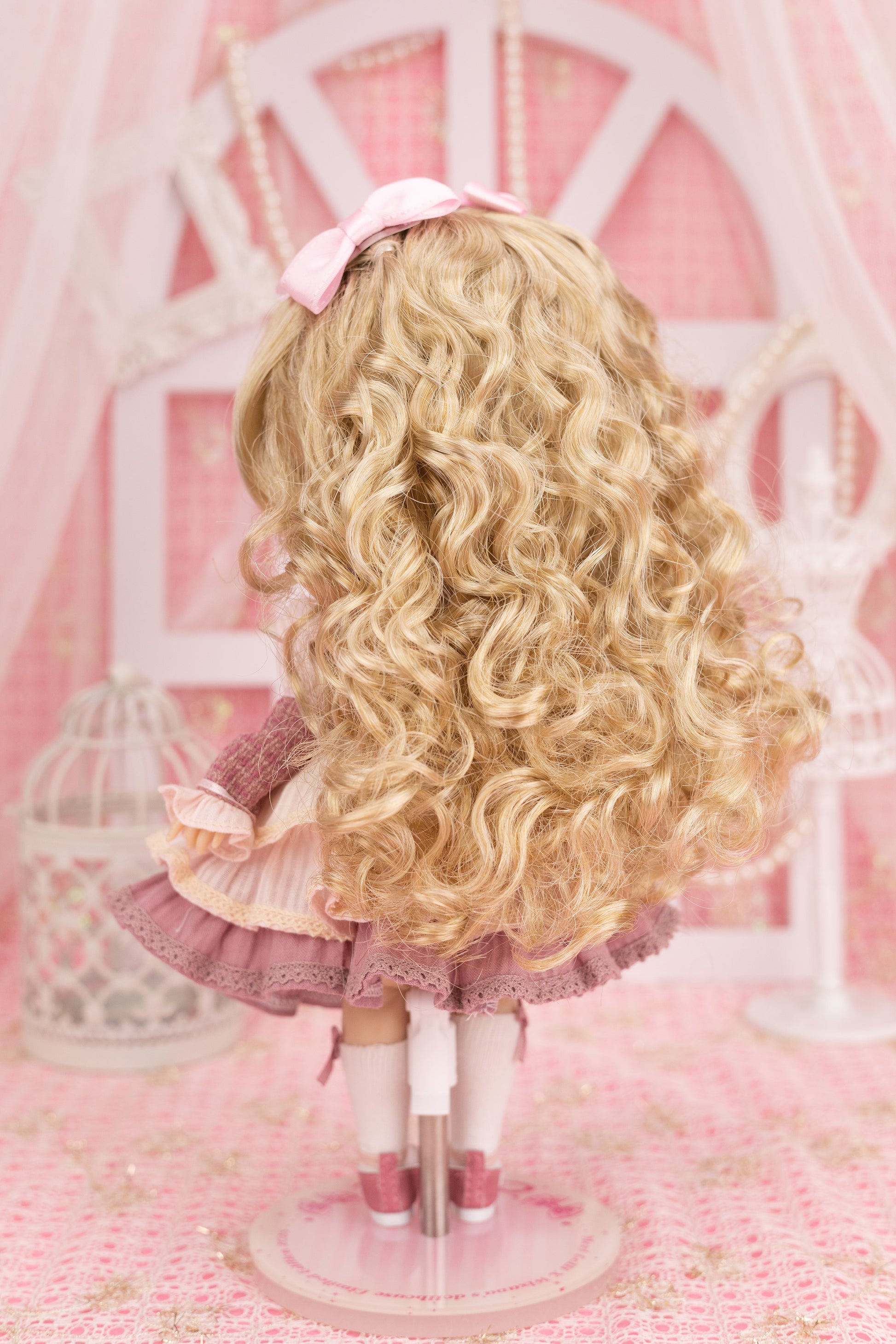 Backview of Candice doll with blonde curls and a pink bow on a pink background