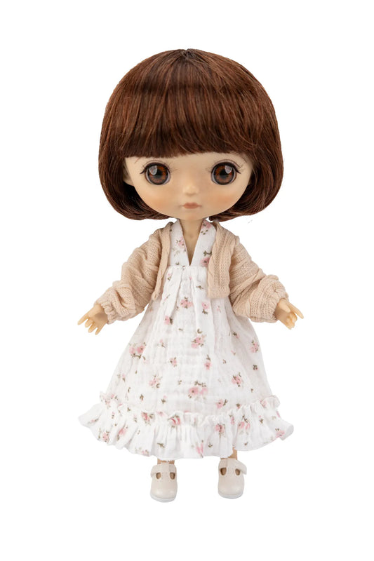 A doll with brown hair and large eyes wearing a white floral dress with a beige cardigan and white shoes, shown against a plain white background.