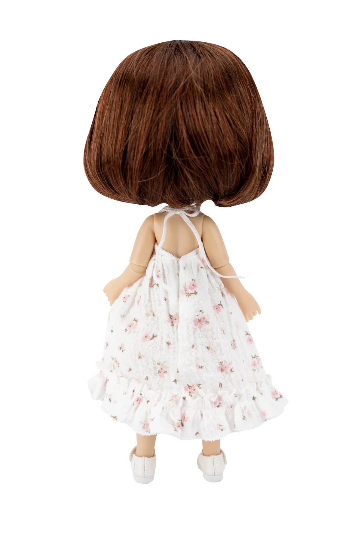 A doll viewed from the back, featuring short brown hair and wearing a white sleeveless ruffled dress with a pink floral pattern and white shoes, isolated on a white background.