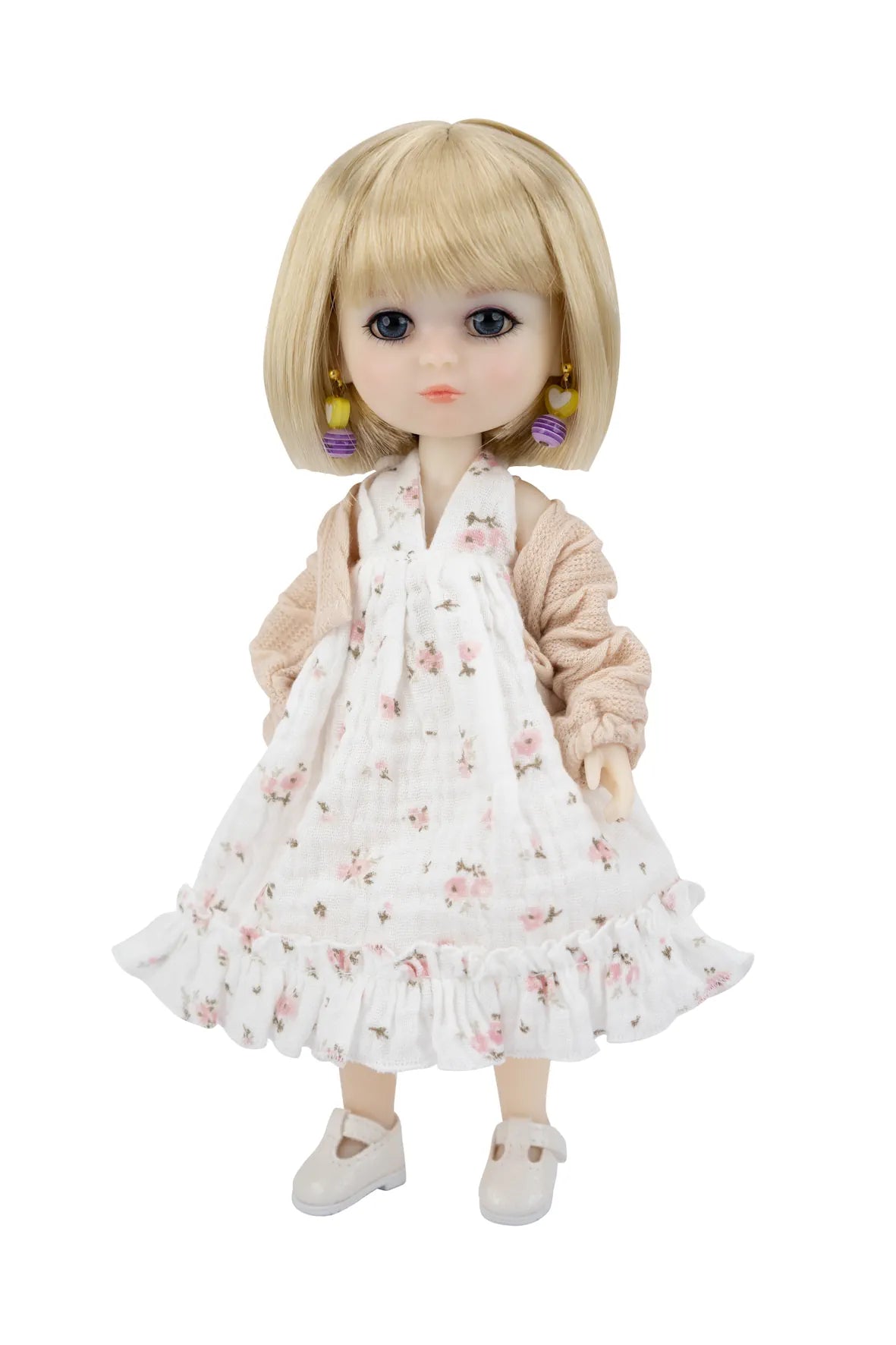 A blonde doll with large dark eyes wearing a white floral pinafore dress, a beige cardigan, and white shoes, shown against a plain white background.