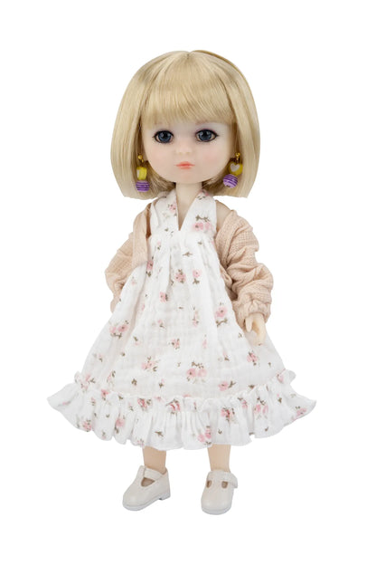 A blonde doll with large dark eyes wearing a white floral pinafore dress, a beige cardigan, and white shoes, shown against a plain white background.
