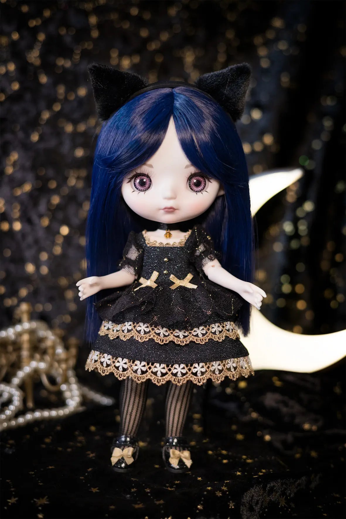 A doll with long dark blue hair, black cat ears, and a black dress with gold lace trim, posed against a dark starry backdrop with a crescent moon and pearl beads.