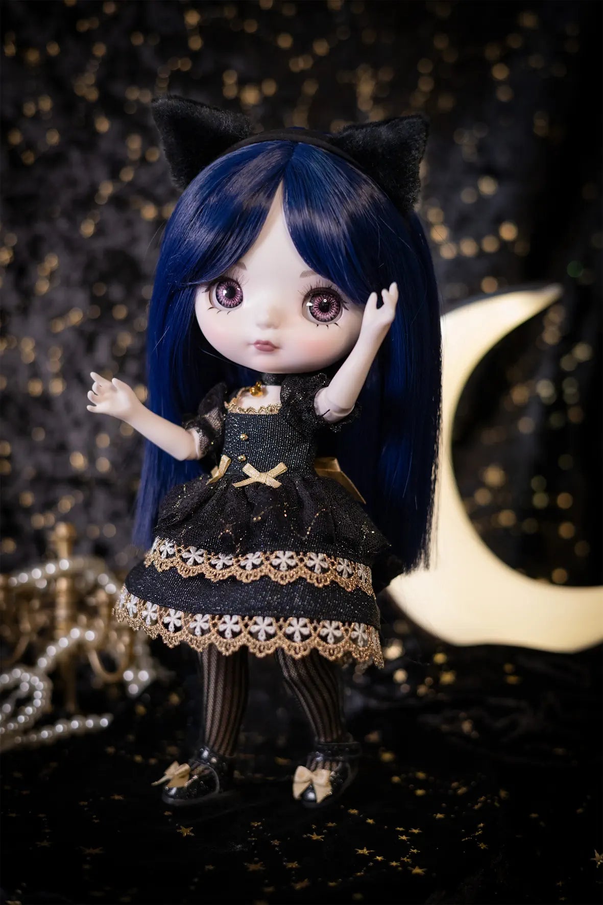 A doll with long dark blue hair, black cat ears, and a black dress with gold lace trim stands against a dark background with gold bokeh lights and a crescent moon prop.