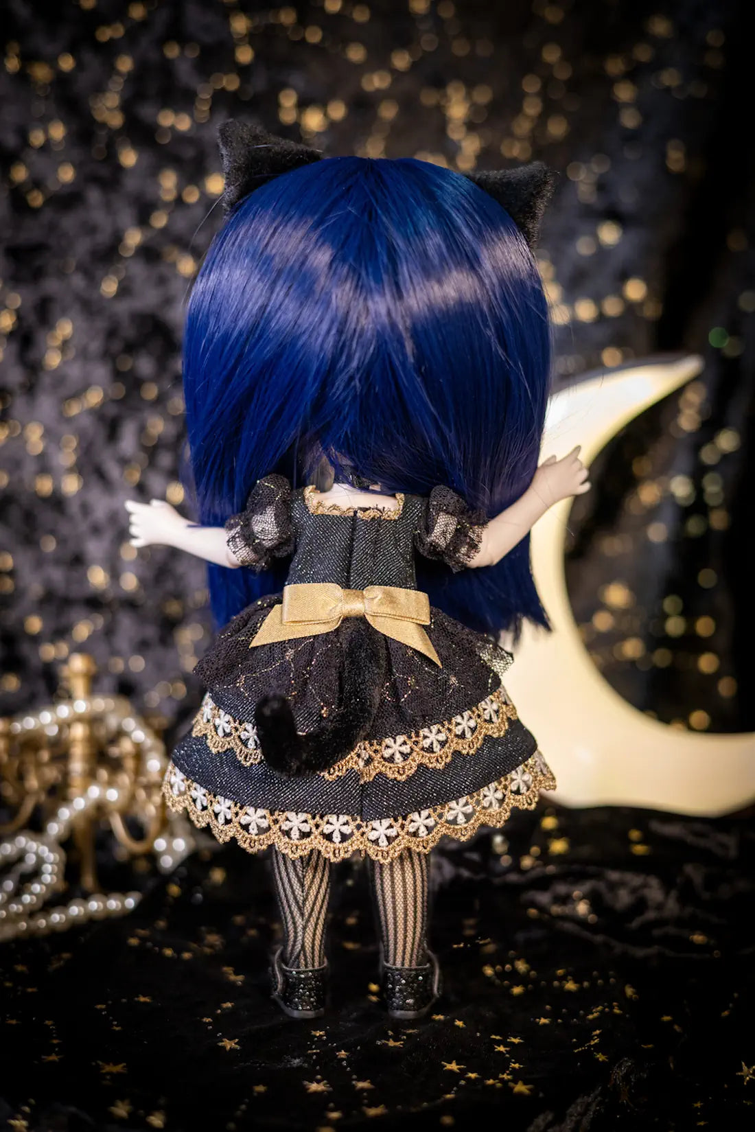 A doll viewed from behind wearing a black and gold gothic-style dress with a large blue wig and black cat ears, posed against a dark starry backdrop with a crescent moon and pearl strands.
