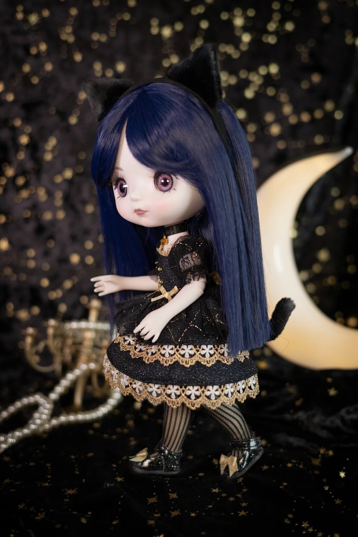 A doll with long dark blue hair, black cat ears, and a black dress with gold lace trim, posed against a dark background with gold bokeh lights and a crescent moon prop.