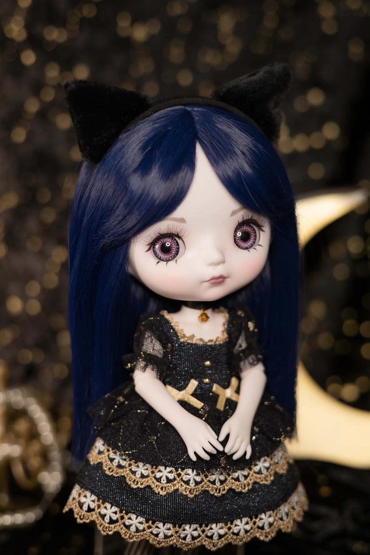 A doll with long dark blue hair, large expressive eyes, and black cat ears, wearing a black dress with gold lace trim and a gold cross detail on the chest, posed against a dark glittery background.