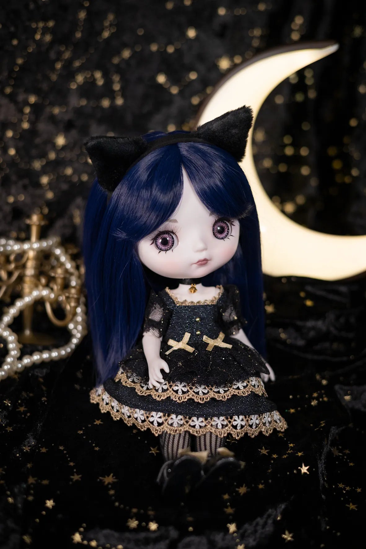 A doll with long dark blue hair, large eyes, and black cat ears, wearing a black dress with gold trim and lace details, posed against a black starry backdrop with a glowing crescent moon.
