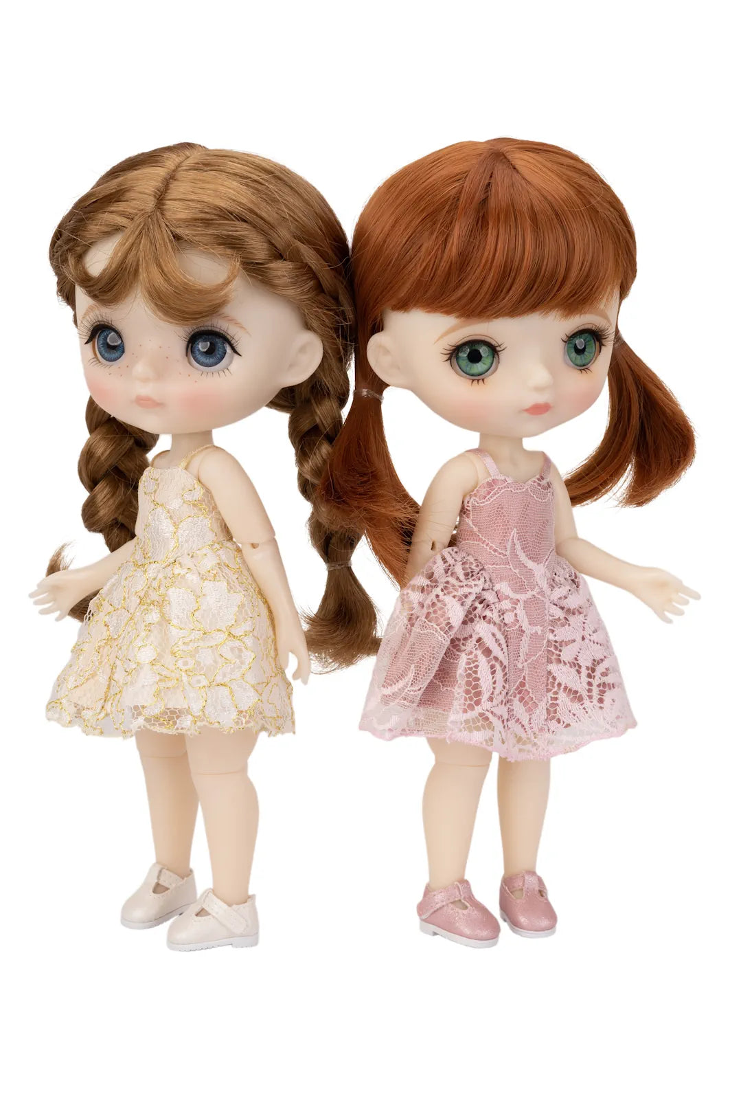 Two Blythe-style dolls with large eyes standing side by side against a white background. The doll on the left has light brown braided hair and wears a cream dress with gold floral pattern and white shoes.