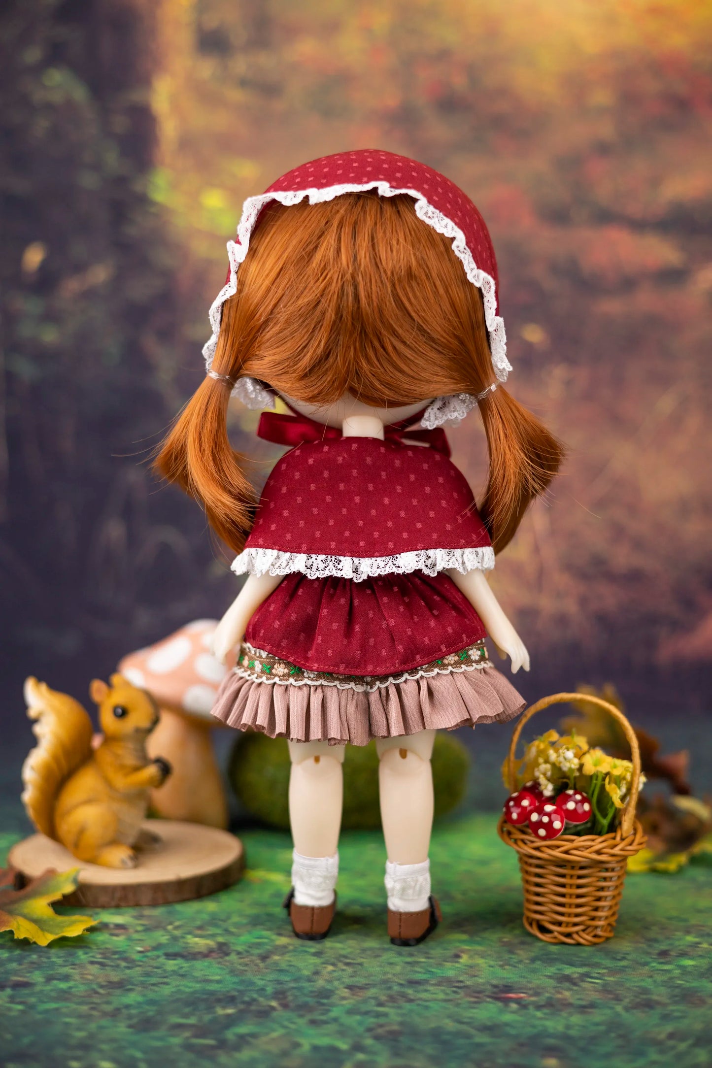 Doll with red dress and bonnet standing in a forest setting with a basket and flowers.
