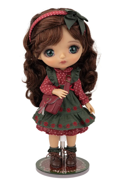 A Mori Lolli doll with long brown wavy hair, large blue eyes, wearing a red and green patterned dress, a matching headband with a bow, and brown shoes, standing on a round display base against a white background.