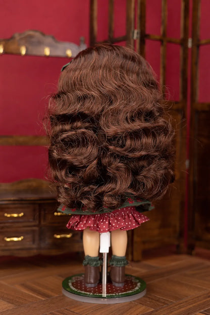 Back view of a doll with long, wavy brown hair, wearing a red polka-dot skirt and dark green boots, standing on a display stand in front of a red wall and wooden furniture.