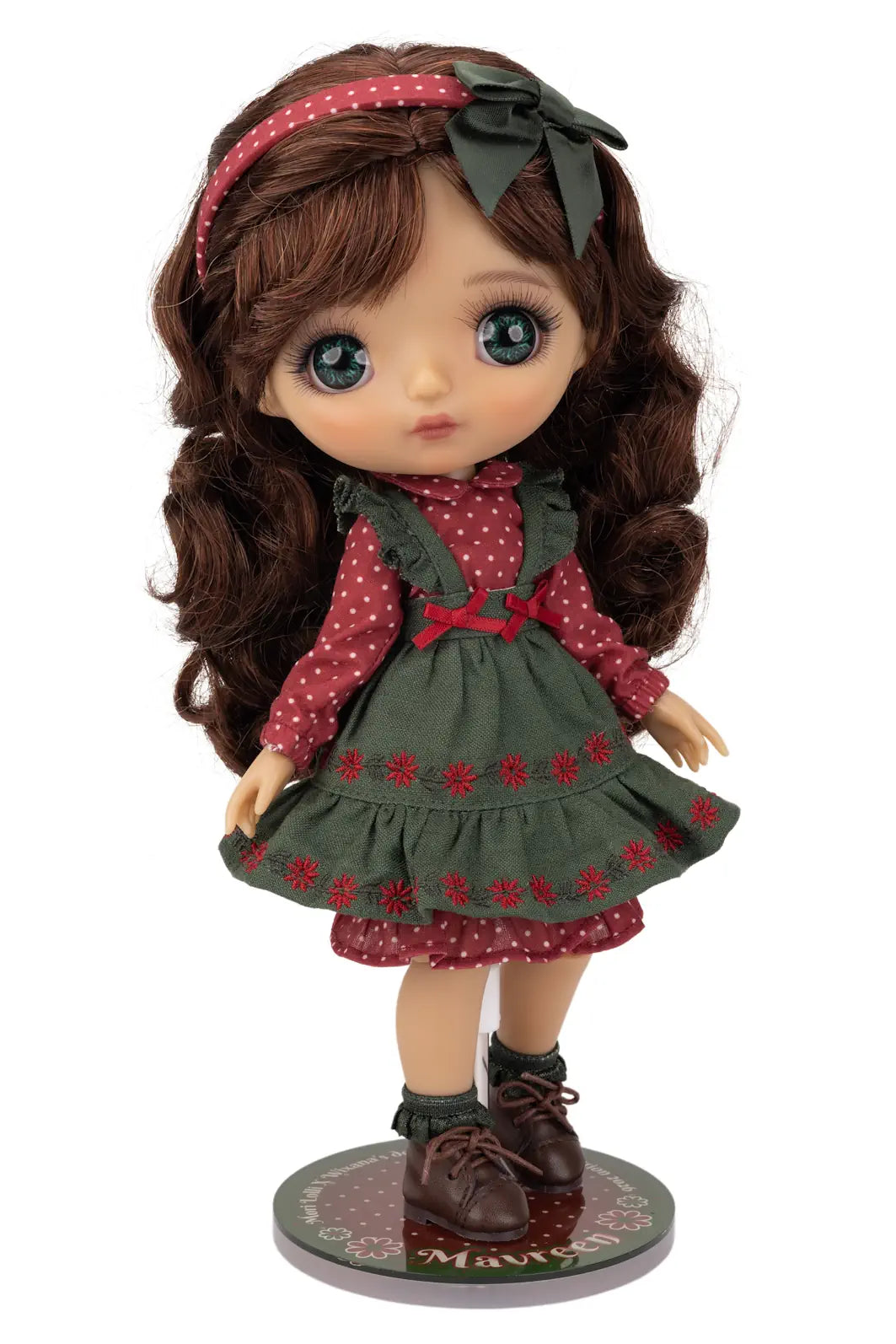 A Mori Lolli doll with long brown wavy hair, large blue eyes, and a red-and-white polka dot dress layered under an olive green pinafore-style dress, standing on a round display base against a white background.