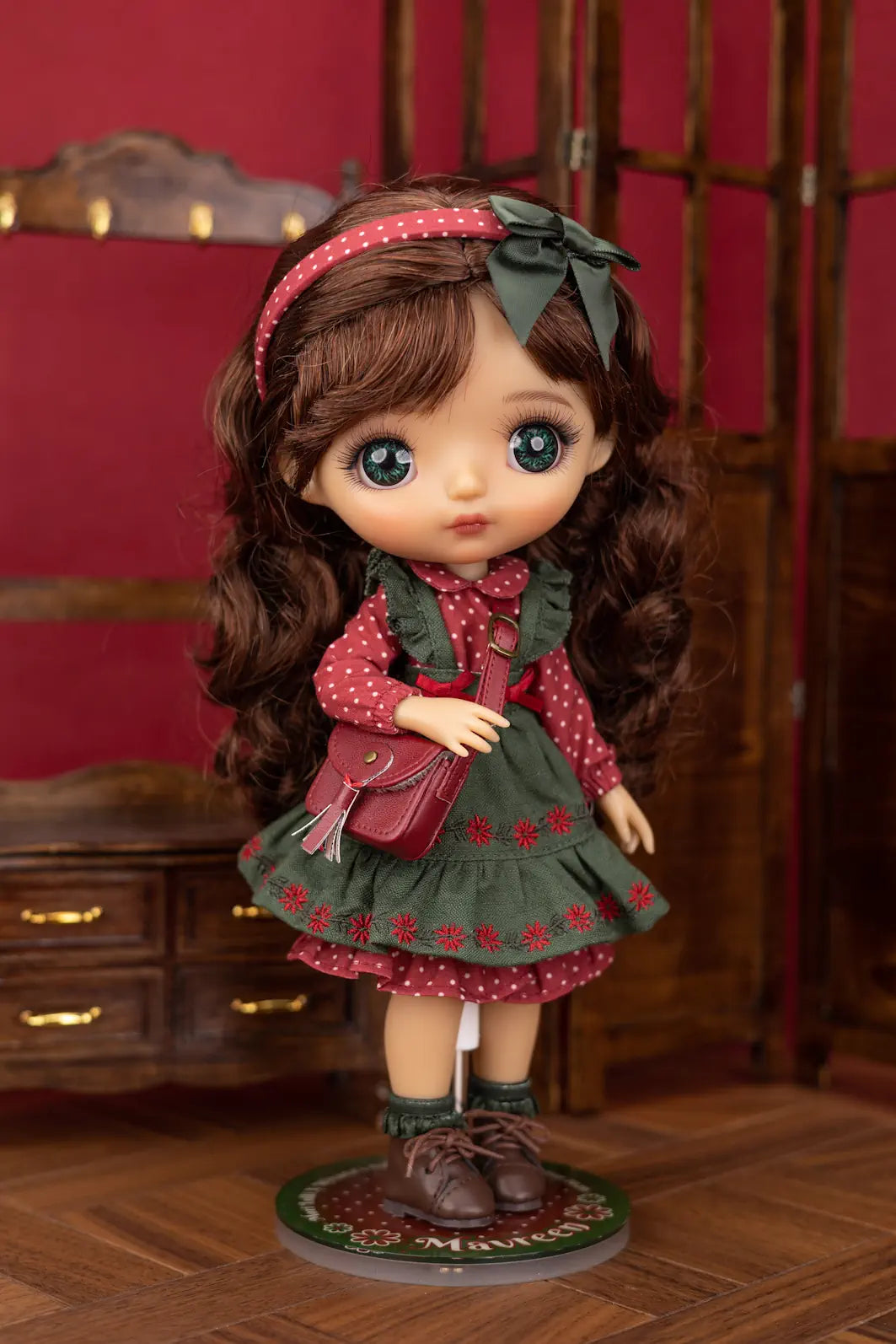 A Mori Lolli doll with long brown wavy hair, large blue eyes, and a green bow headband, wearing a red and green patterned dress and holding a small red bag, posed on a wooden floor against a dark red and brown background.