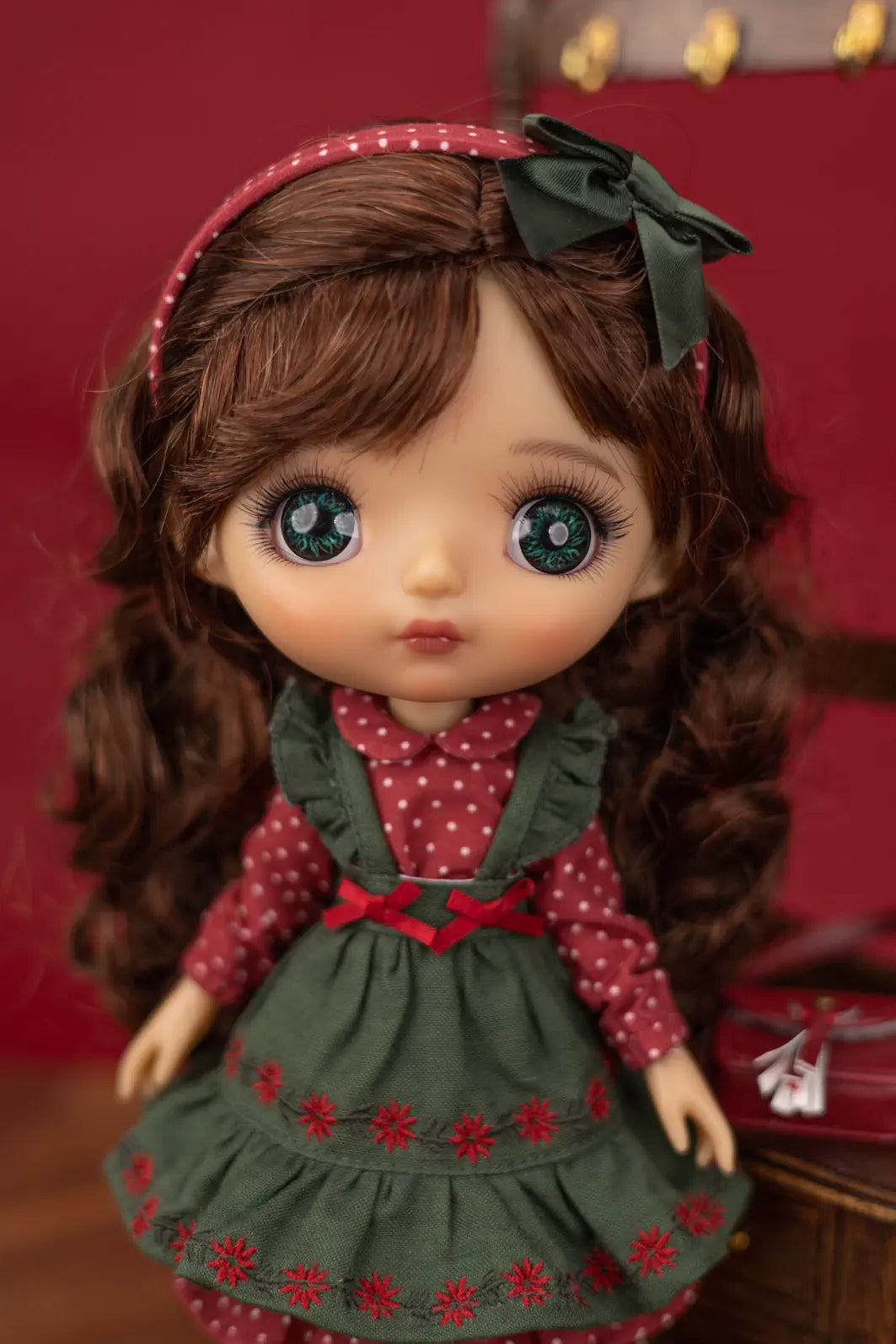 A close-up of a  Mori Lolli doll with large blue eyes, brown hair, and a green bow, wearing a red and green dress against a dark red background.