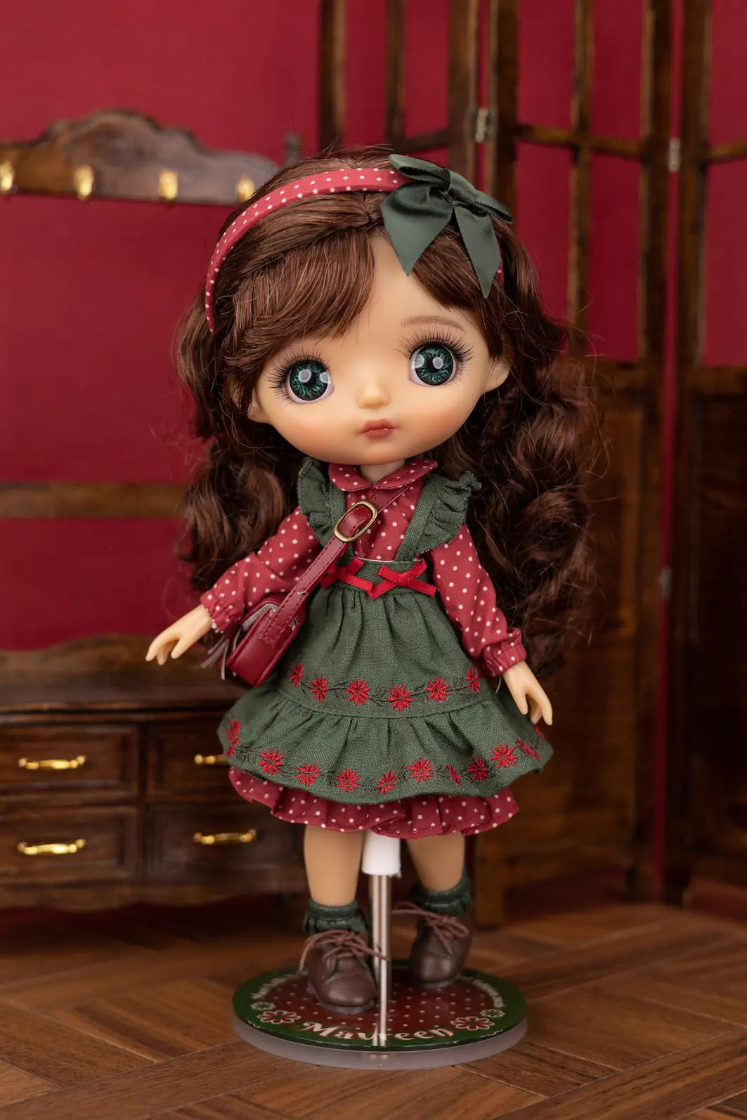 A Mori Lolli doll with long brown hair and large blue eyes, wearing a red and green polka-dot dress, green bow headband, and brown shoes, standing on a display stand in front of a dark wooden cabinet and red wall.