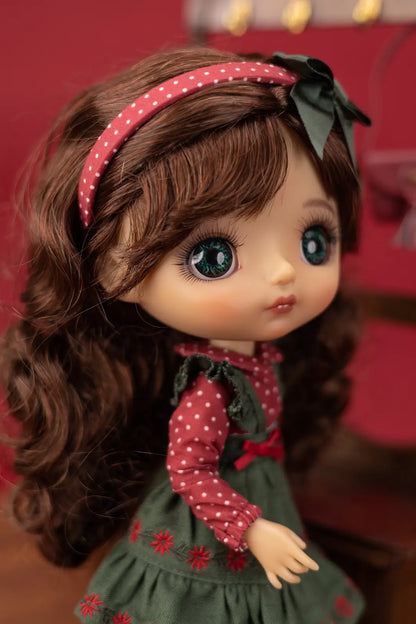 A close-up of a doll with long brown wavy hair, wearing a red polka-dot headband with a dark green bow, a red long-sleeve top with white polka dots, and a dark green dress with red floral embroidery.