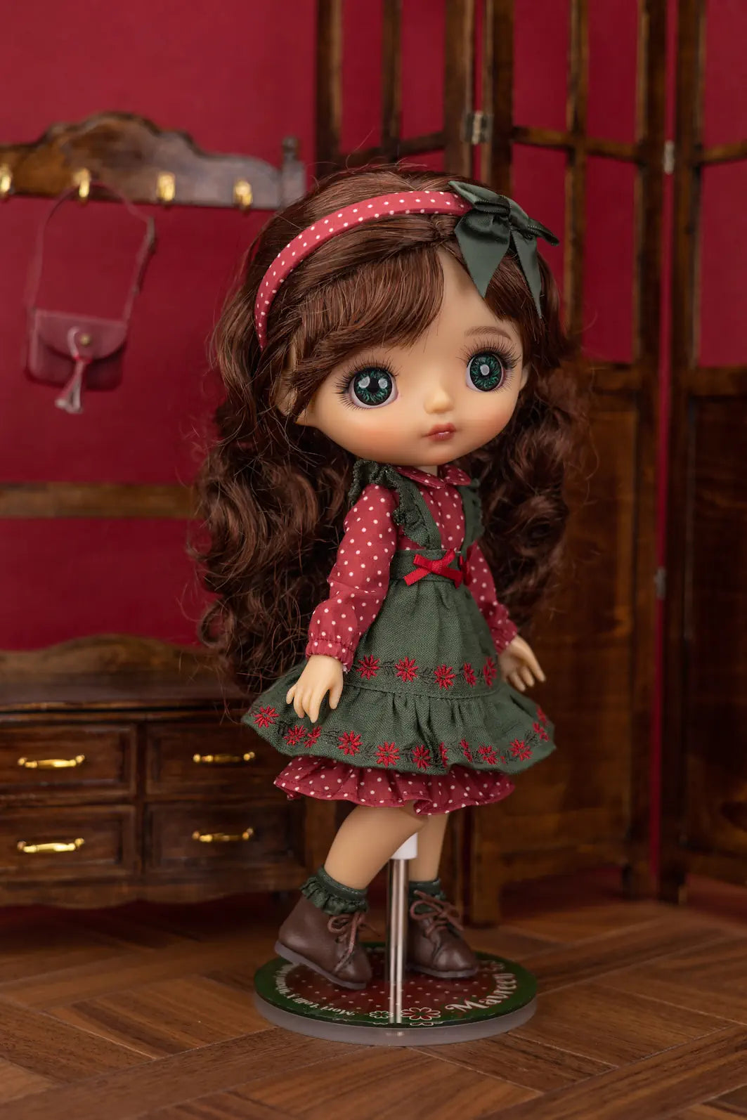 A Mori Lolli doll with long brown curly hair, large blue eyes, and a pink polka-dot headband, wearing a green dress with red polka-dot sleeves and red floral accents, standing on a display stand in a dollhouse-style room with red walls and wooden furniture.