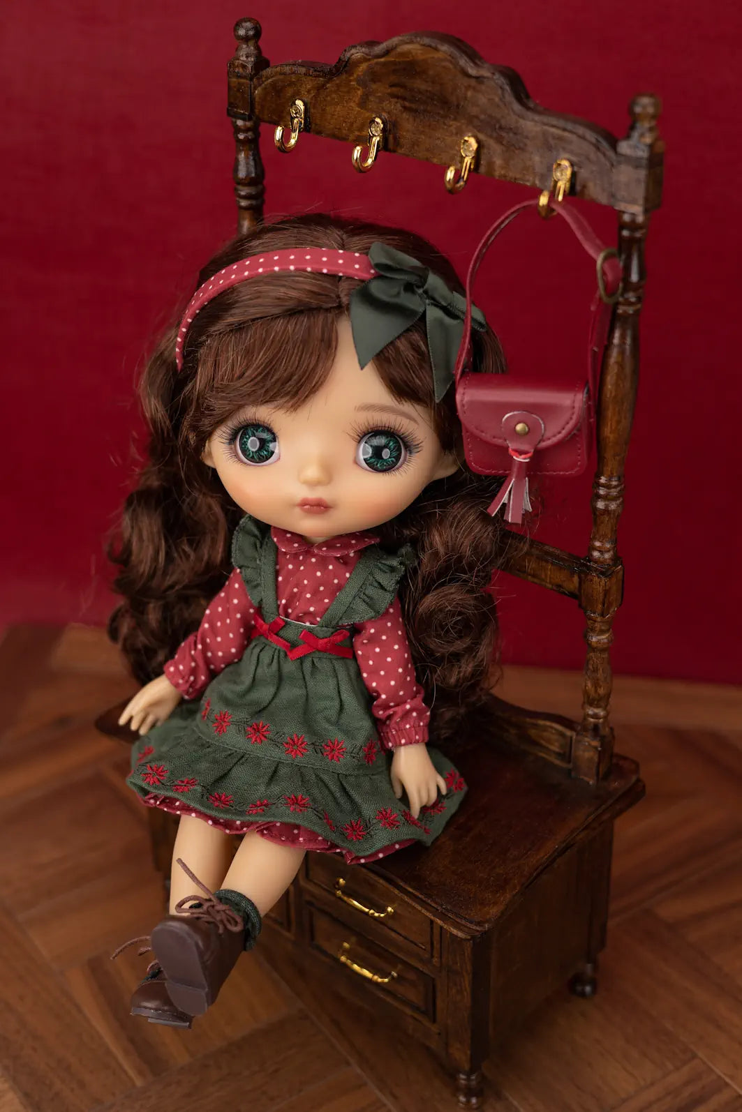 A doll with long brown hair and large blue eyes sits on a miniature wooden chair, wearing a green and red polka-dot dress, a dark green bow headband, and dark brown shoes. A small pink purse hangs from the chair against a dark red background.