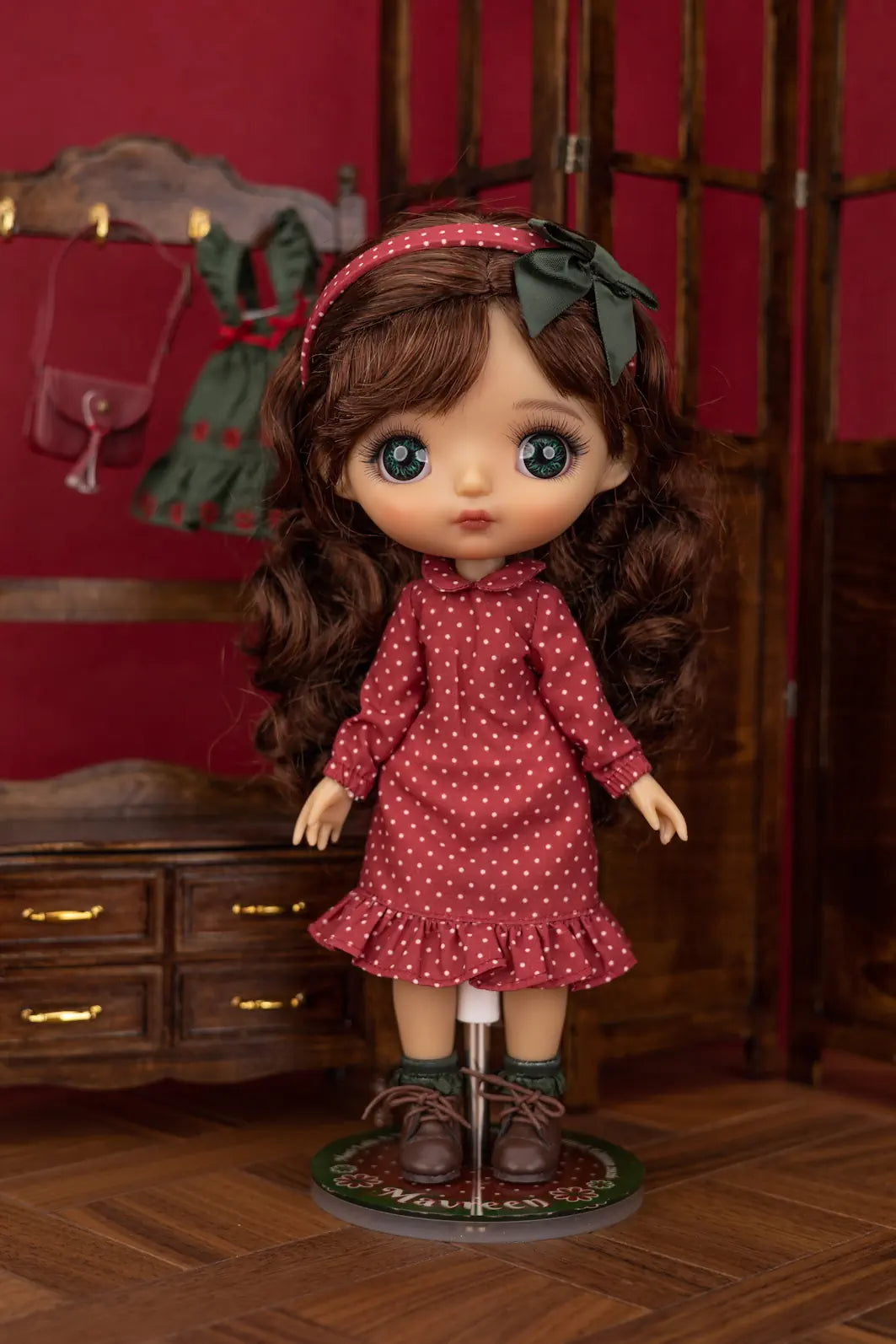 A doll with long brown curly hair, large blue eyes, and a red polka-dot dress, standing on a display stand in a dollhouse-style room with a red wall and wooden furniture.