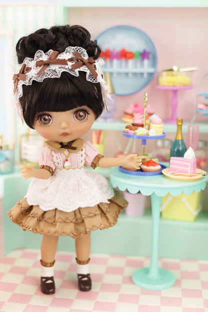Doll in a vintage outfit with a miniature birthday cake and decorations in a pastel-colored room.