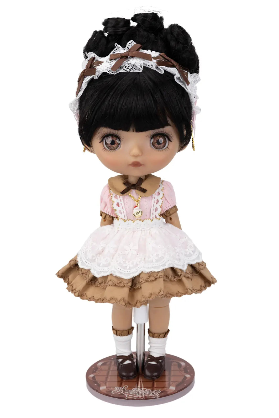 A doll with large brown eyes and black hair styled in a messy bun with a lace headband, wearing a pink and brown ruffled dress with white lace trim, standing on a round display base against a white background.