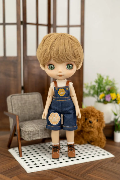 A Mori Lolli doll with light brown hair and green eyes, wearing blue denim overalls with a paw-print patch on the leg, standing on a miniature black-and-white checkered rug in a dollhouse-style room with a gray armchair, wooden floor, and a small plush dog.