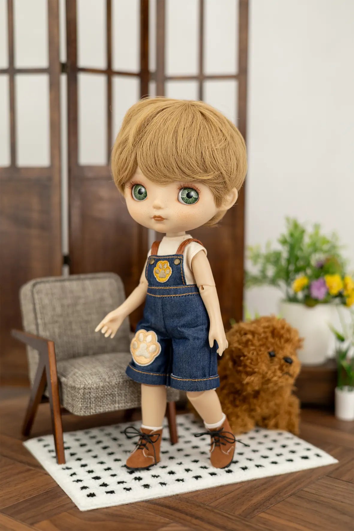 A doll with blonde hair and green eyes wearing blue denim-style overalls with a paw-print patch, standing on a patterned rug in a miniature room setting with a chair, teddy bear, and plants.