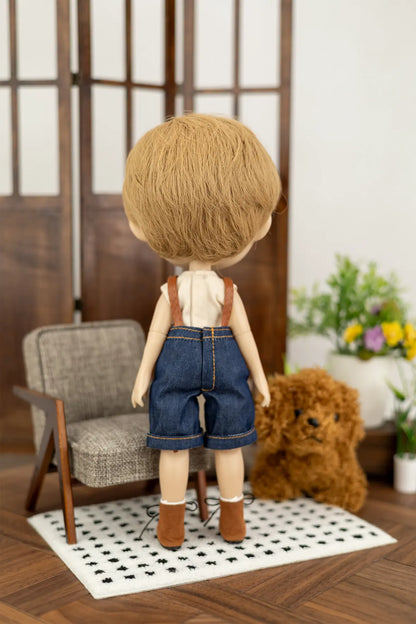 A doll viewed from behind wearing a beige tank top and blue denim shorts, standing on a small rug in a miniature room setup with a chair, rug, and plush toy.