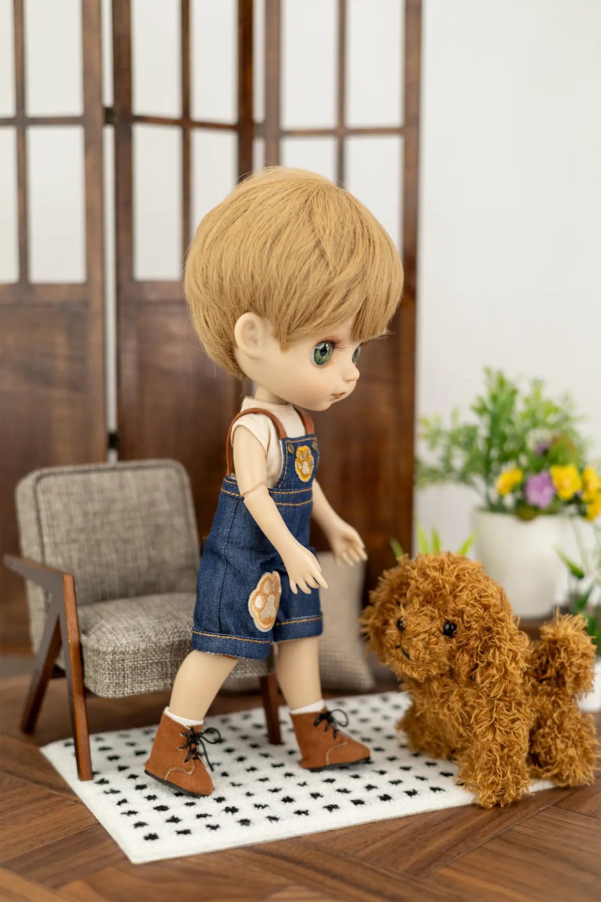 A doll with short blonde hair, wearing blue denim overalls and brown shoes, standing on a black-and-white checkered rug next to a small brown plush dog in a miniature room setting.