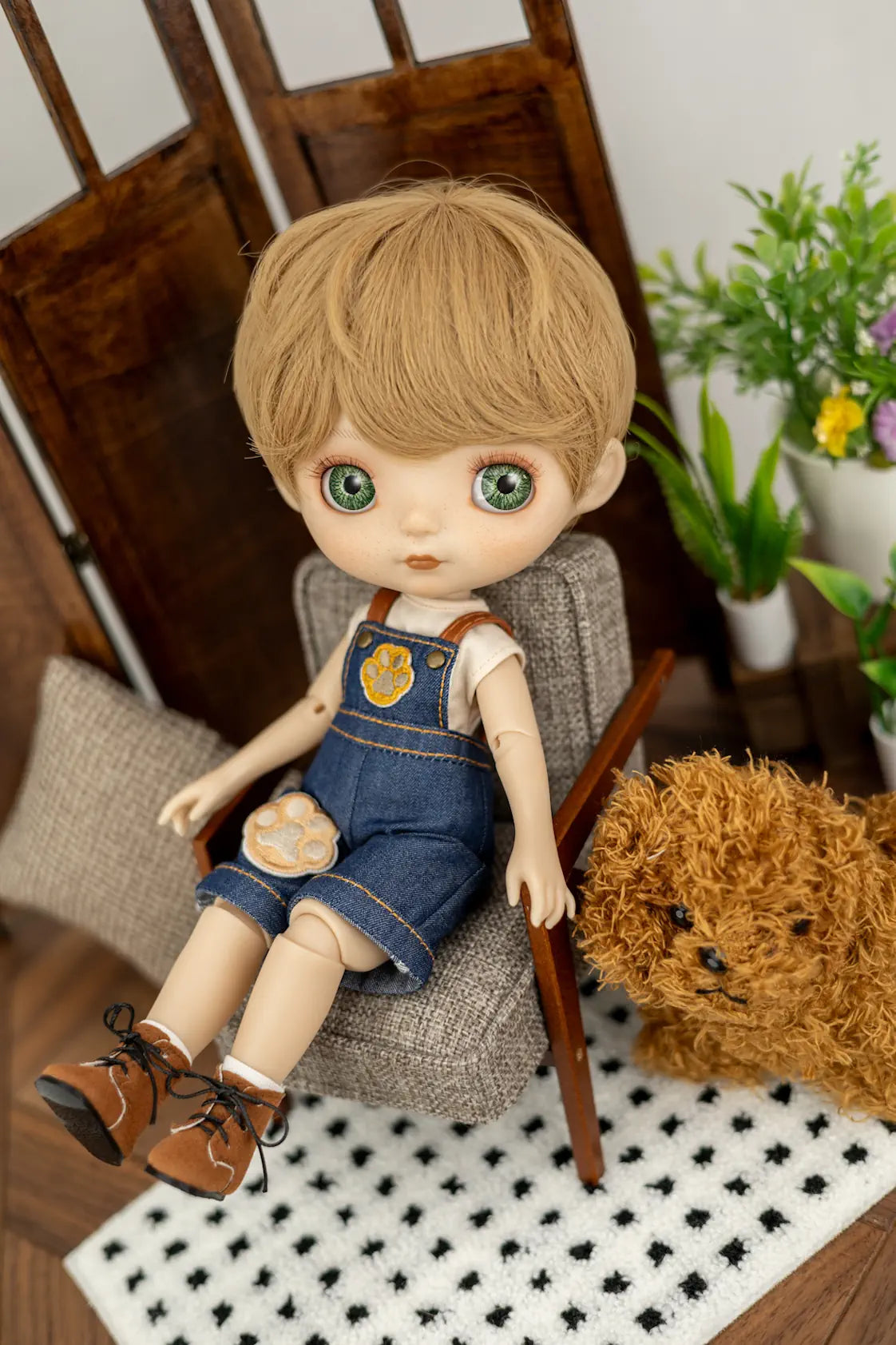 A doll with blonde hair and green eyes sits in a miniature gray armchair, wearing denim overalls over a white t-shirt and brown shoes. The doll is posed in a miniature room setting with a wooden chair, a small potted plant, and a teddy bear.
