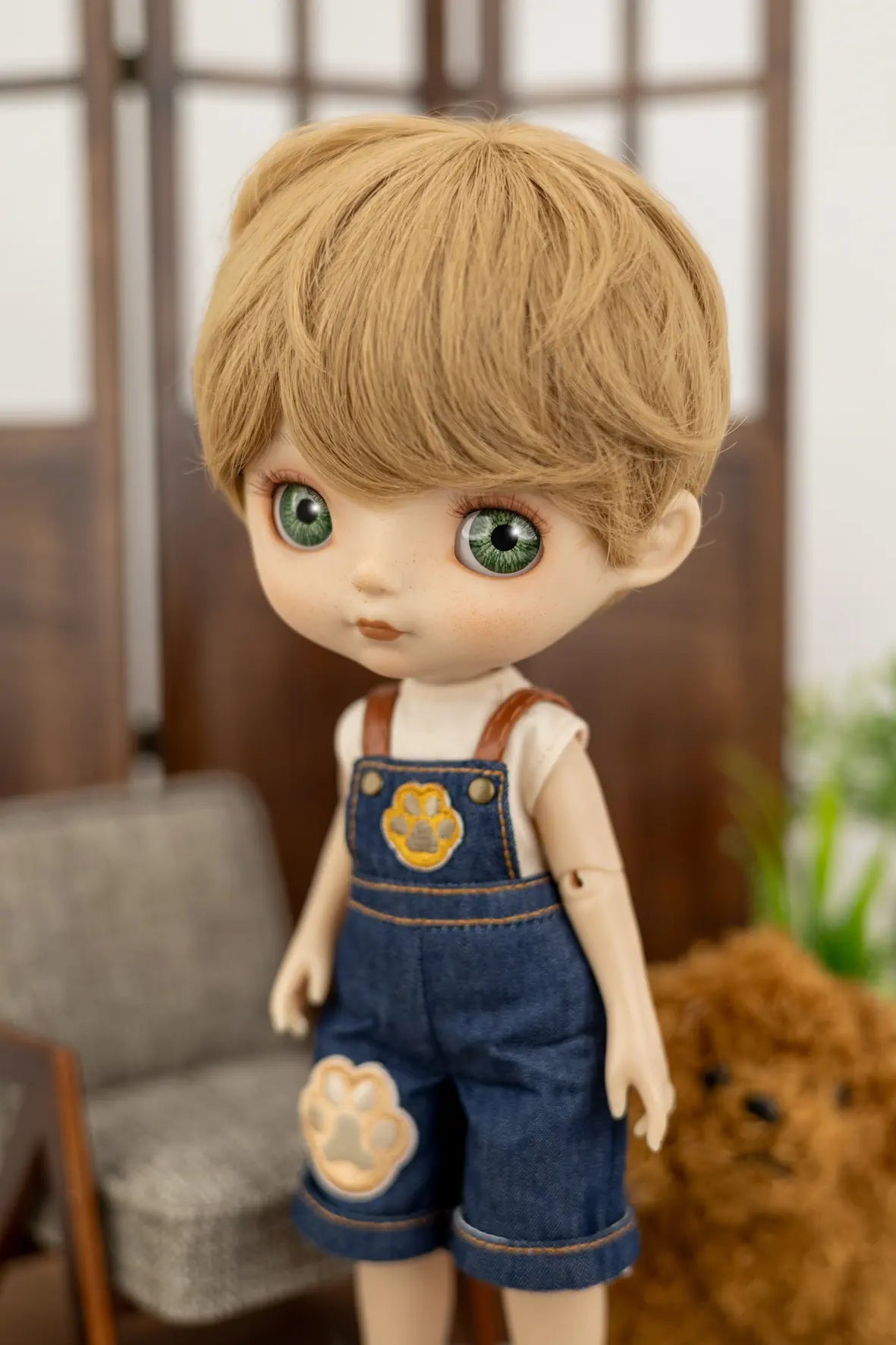 A doll with blonde hair and large green eyes, wearing blue denim-style overalls with a paw-print patch and a white shirt, posed indoors against a softly blurred background.