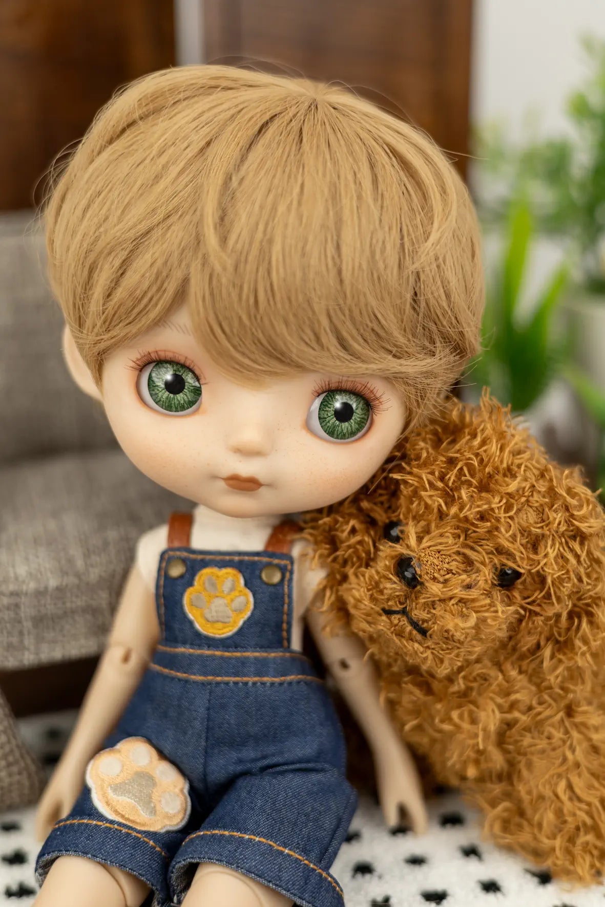 A close-up of a doll with large green eyes, blonde hair, and blue denim overalls, sitting next to a brown curly-haired plush toy.
