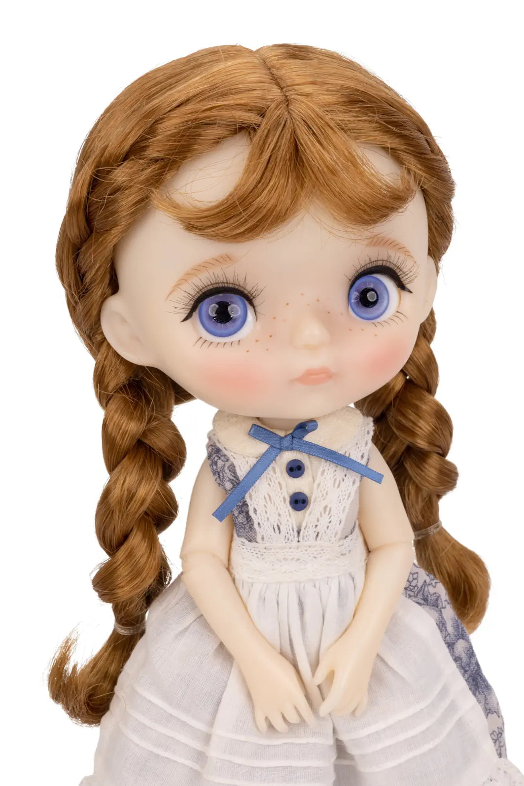 A close-up of a doll with large blue eyes, reddish-brown braided hair, and a white dress with a blue bow, shown against a plain white background.