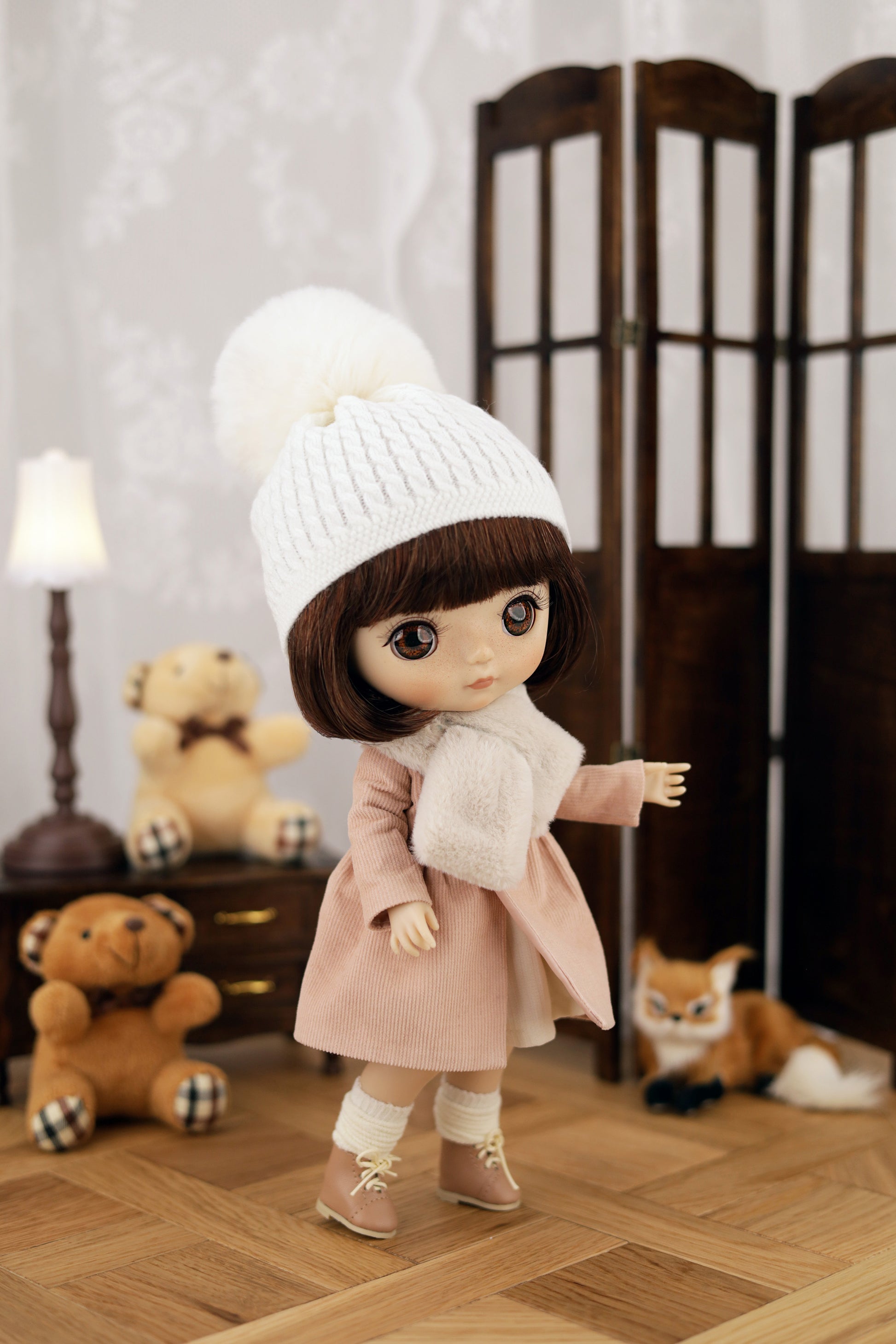 Doll wearing a white knit hat and pink coat in a room with teddy bears and a lamp.
