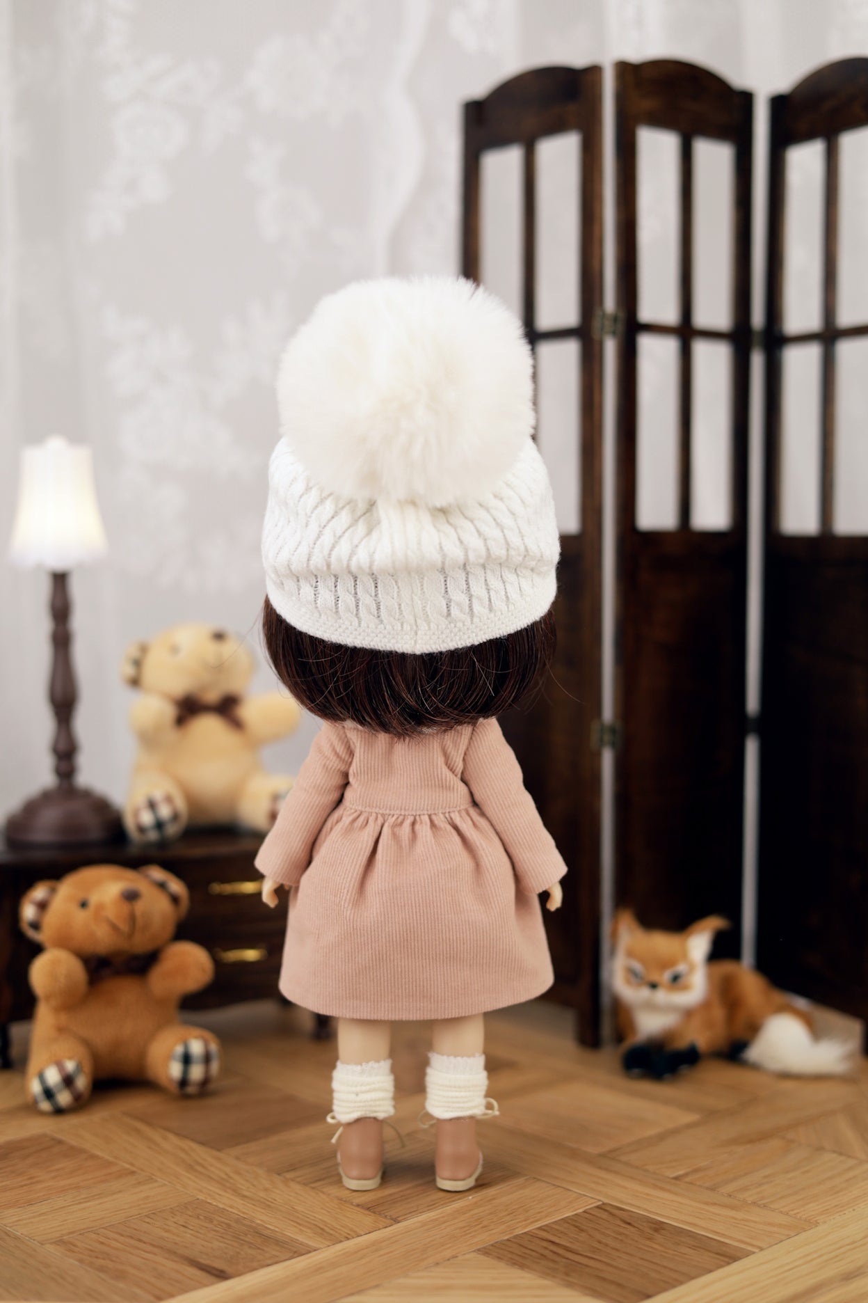 Doll wearing a white beanie and pink dress in a room with teddy bears and a lamp.
